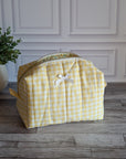 A yellow and white gingham quilted toiletry bag with a small white bow on the front, set on a gray wood floor against a white paneled wall with a potted plant on the left.
