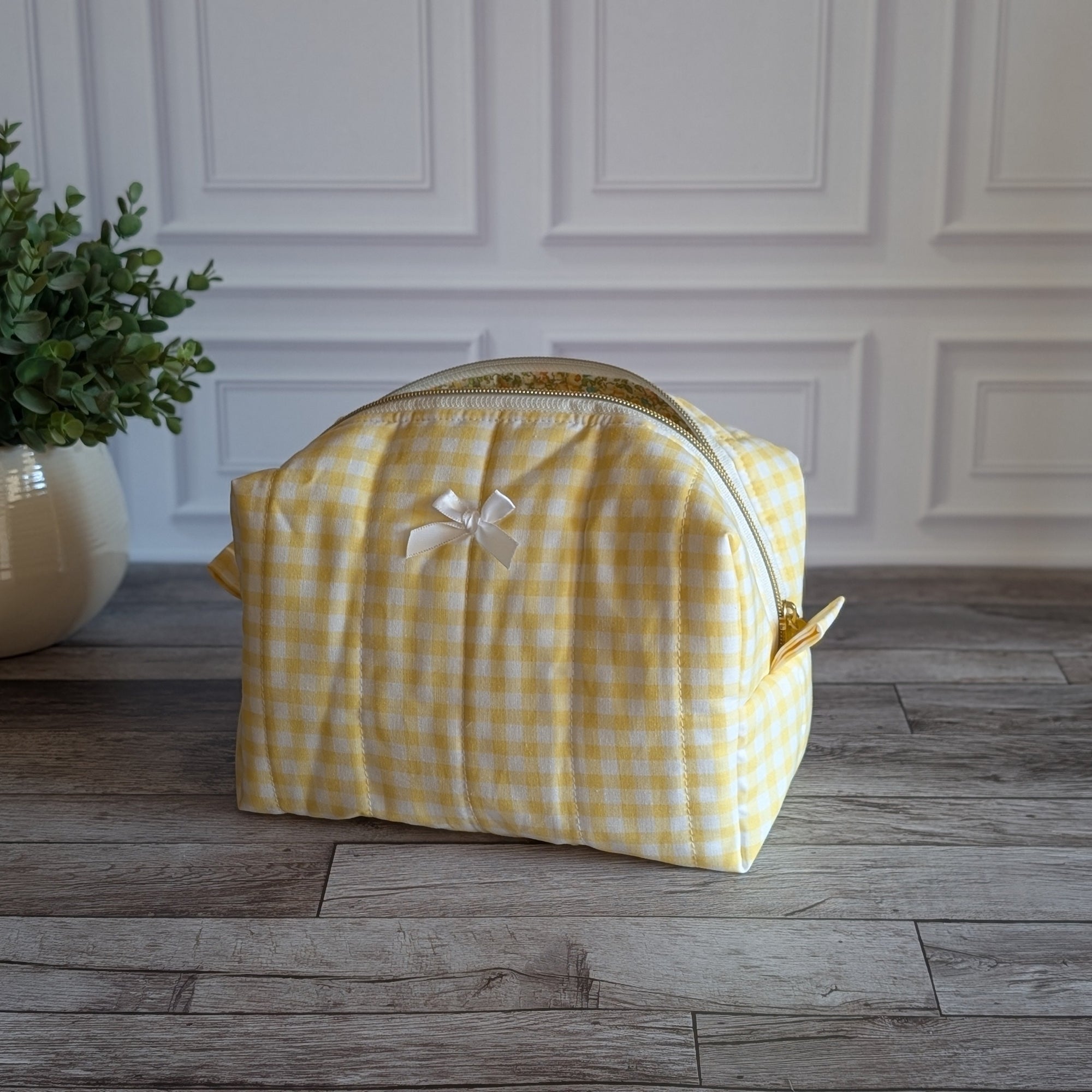 A yellow and white gingham toiletry bag with a small white bow on the front, sitting on a gray wood floor in front of a white paneled wall next to a potted green plant.