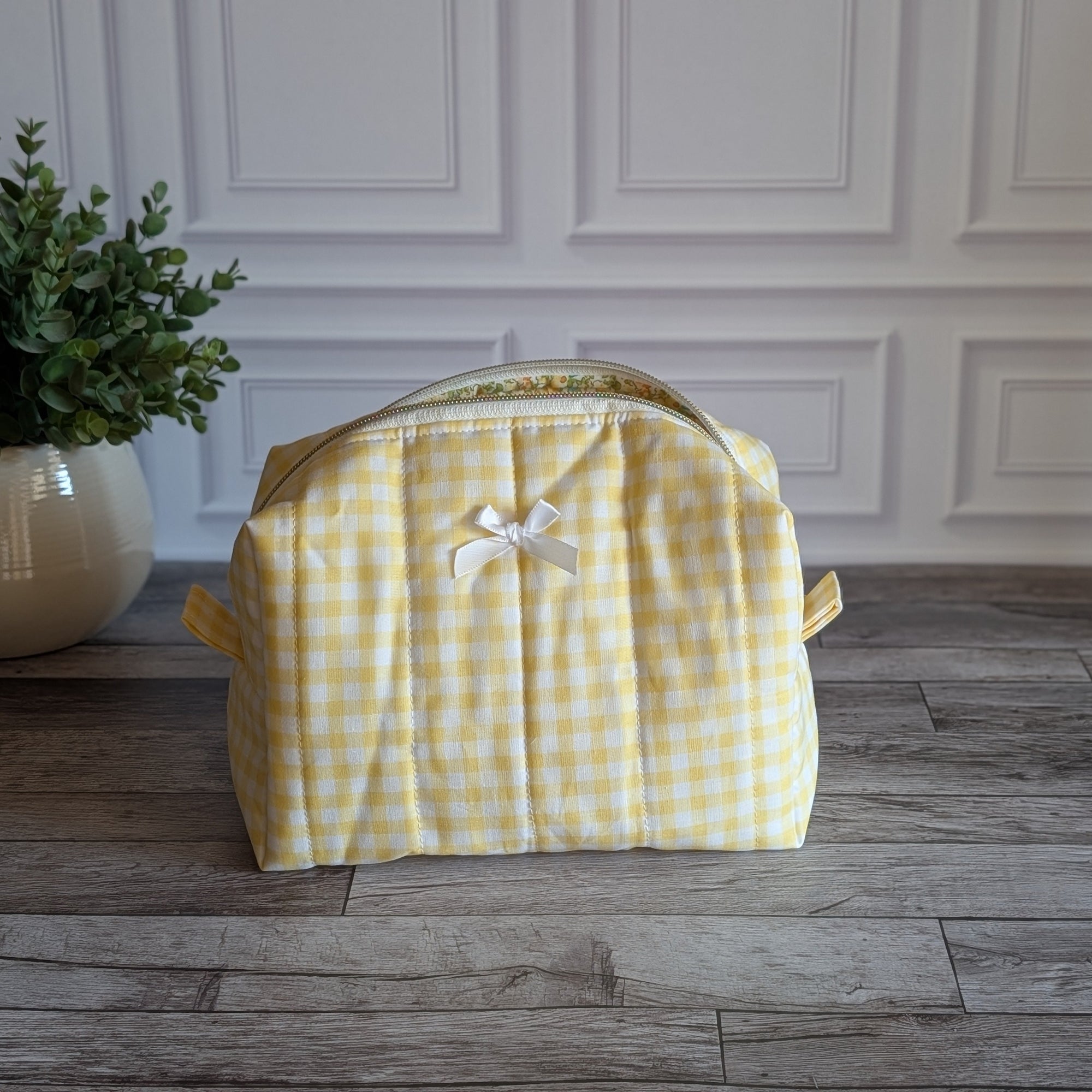 A yellow and white gingham toiletry bag with a small white bow on the front, sitting on a gray wood floor in front of a white paneled wall with a potted plant on the left.