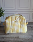 A yellow and white gingham toiletry bag with a small white bow on the front, sitting on a gray wood floor in front of a white paneled wall with a potted plant on the left.