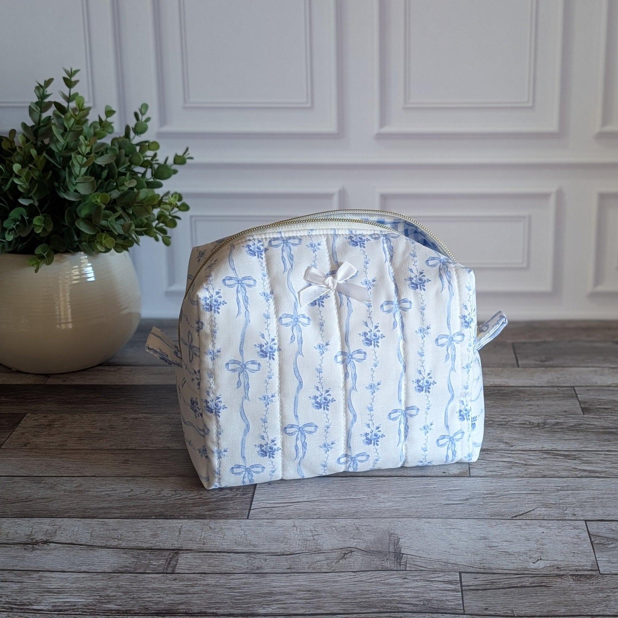A quilted fabric toiletry bag with a blue floral and bow pattern on a white background, sitting on a gray wood floor next to a potted green plant.