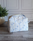 A quilted fabric toiletry bag with a blue floral and bow pattern on a white background, sitting on a gray wood floor next to a potted green plant.