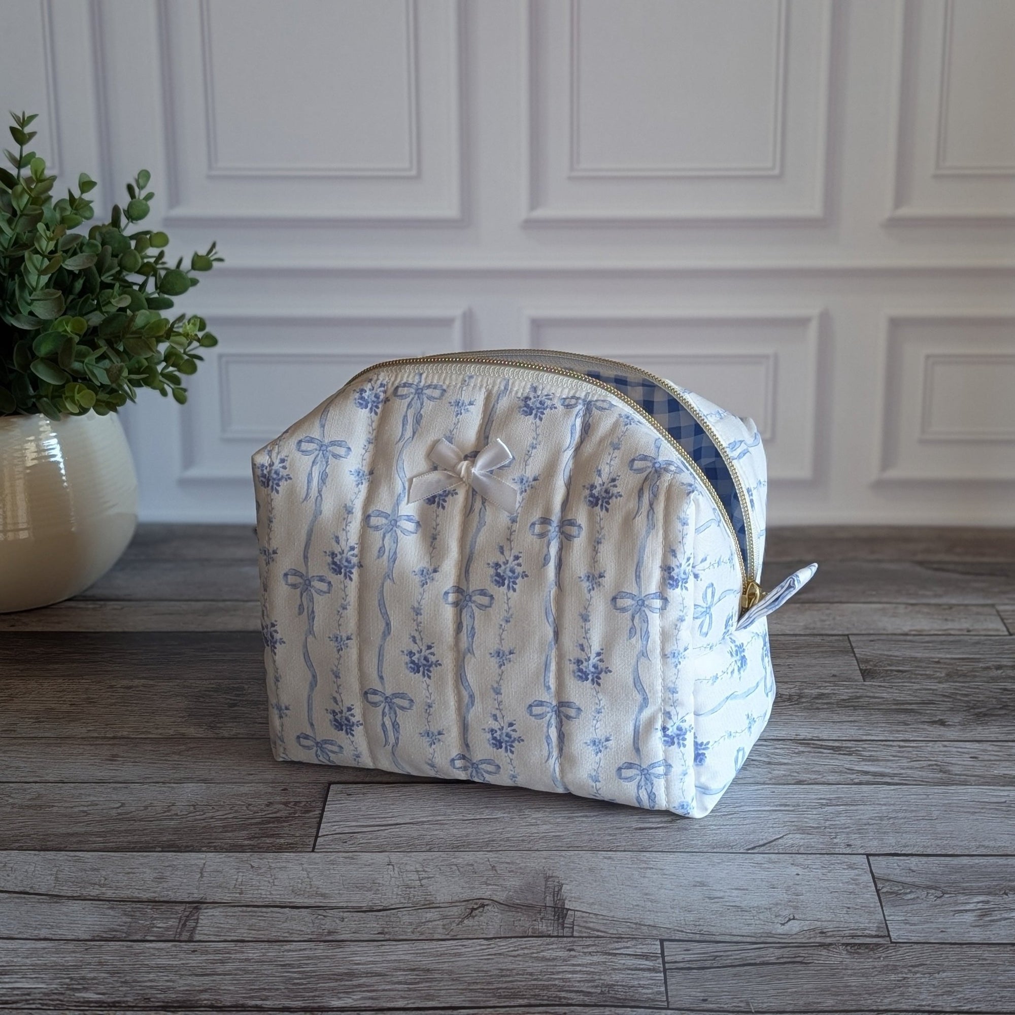 A quilted cosmetic pouch with a blue floral pattern on a cream background, sitting on a gray wood floor in front of a white paneled wall.