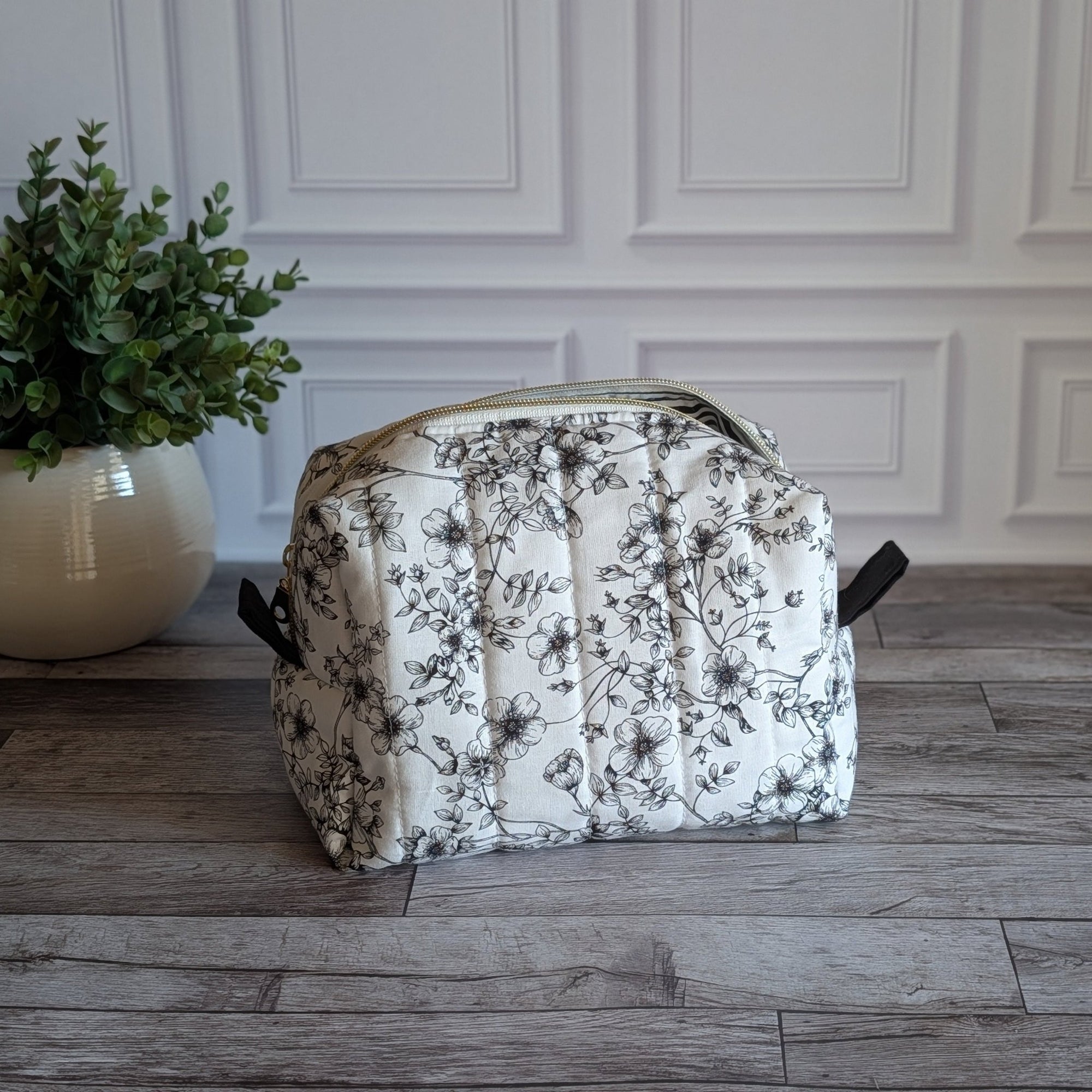 A toiletry bag with a black floral pattern on a white background, sitting on a gray wood floor next to a potted green plant.