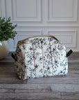 A toiletry bag with a black floral pattern on a white background, sitting on a gray wood floor next to a potted green plant.