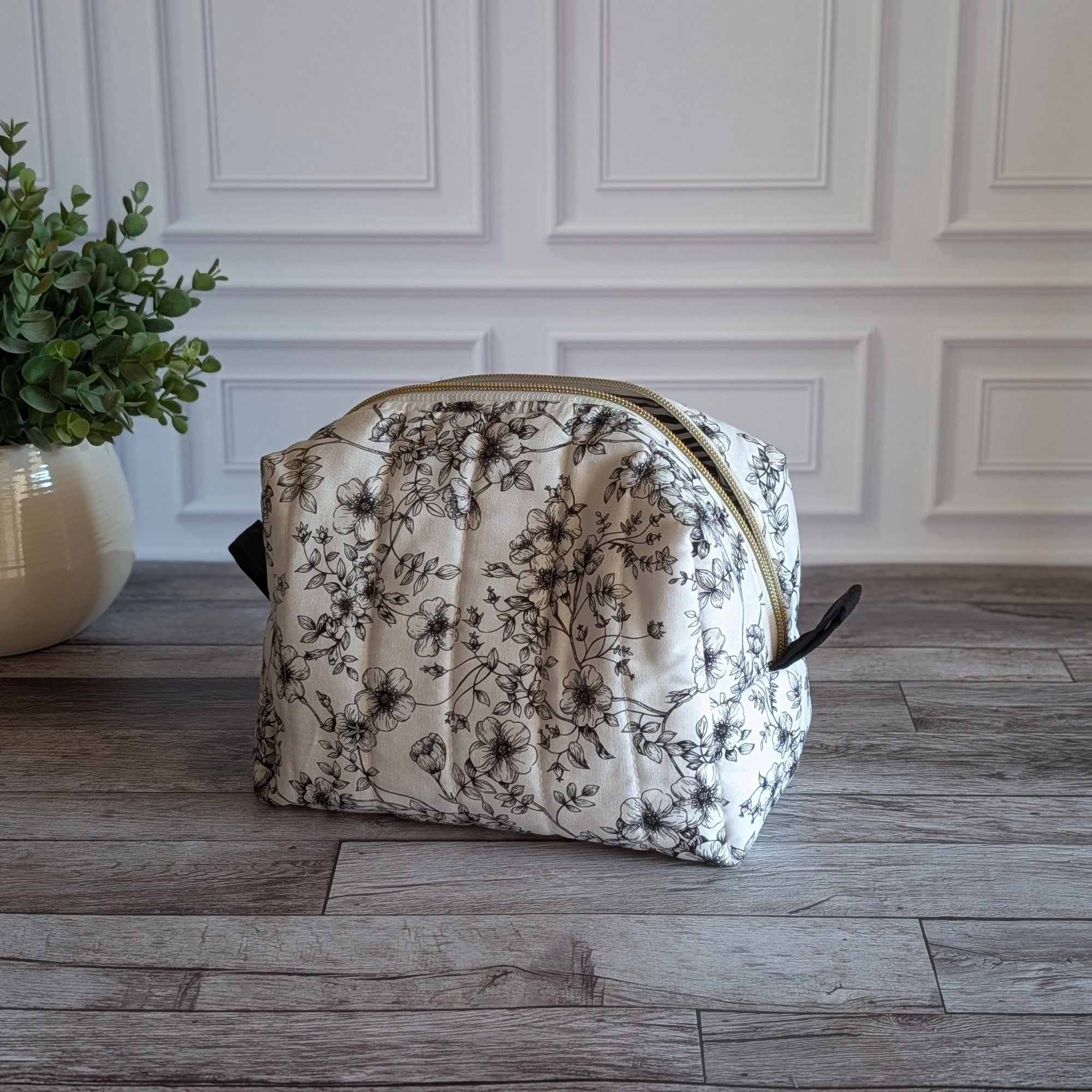 A quilted cosmetic pouch with a black floral toilett pattern on a cream background, featuring a gold zipper and black side tabs, set on a gray wood floor next to a potted green plant.