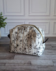 A quilted cosmetic pouch with a black floral toilett pattern on a cream background, featuring a gold zipper and black side tabs, set on a gray wood floor next to a potted green plant.