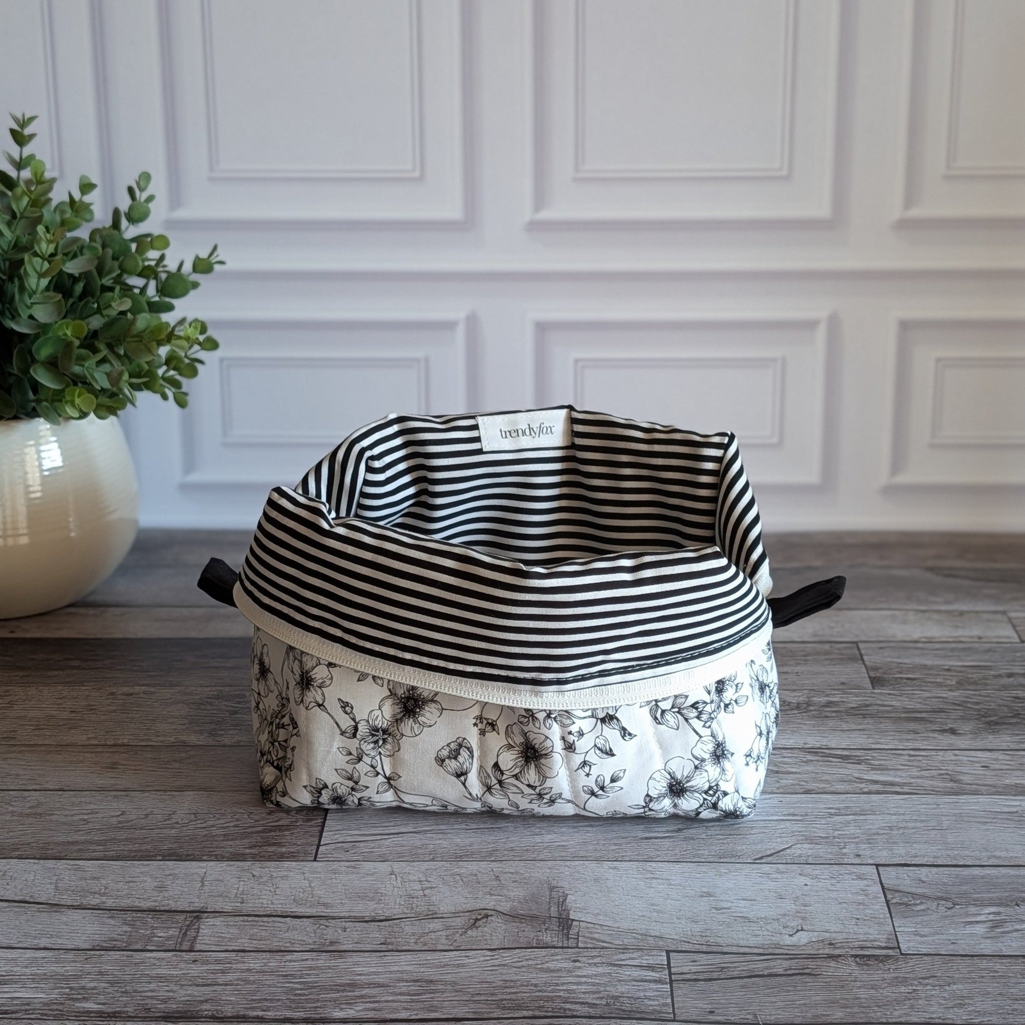 A fabric quilted toiletry with a black and white striped lining and a black-and-white floral patterned outer, placed on a gray wood floor in front of a white paneled wall.