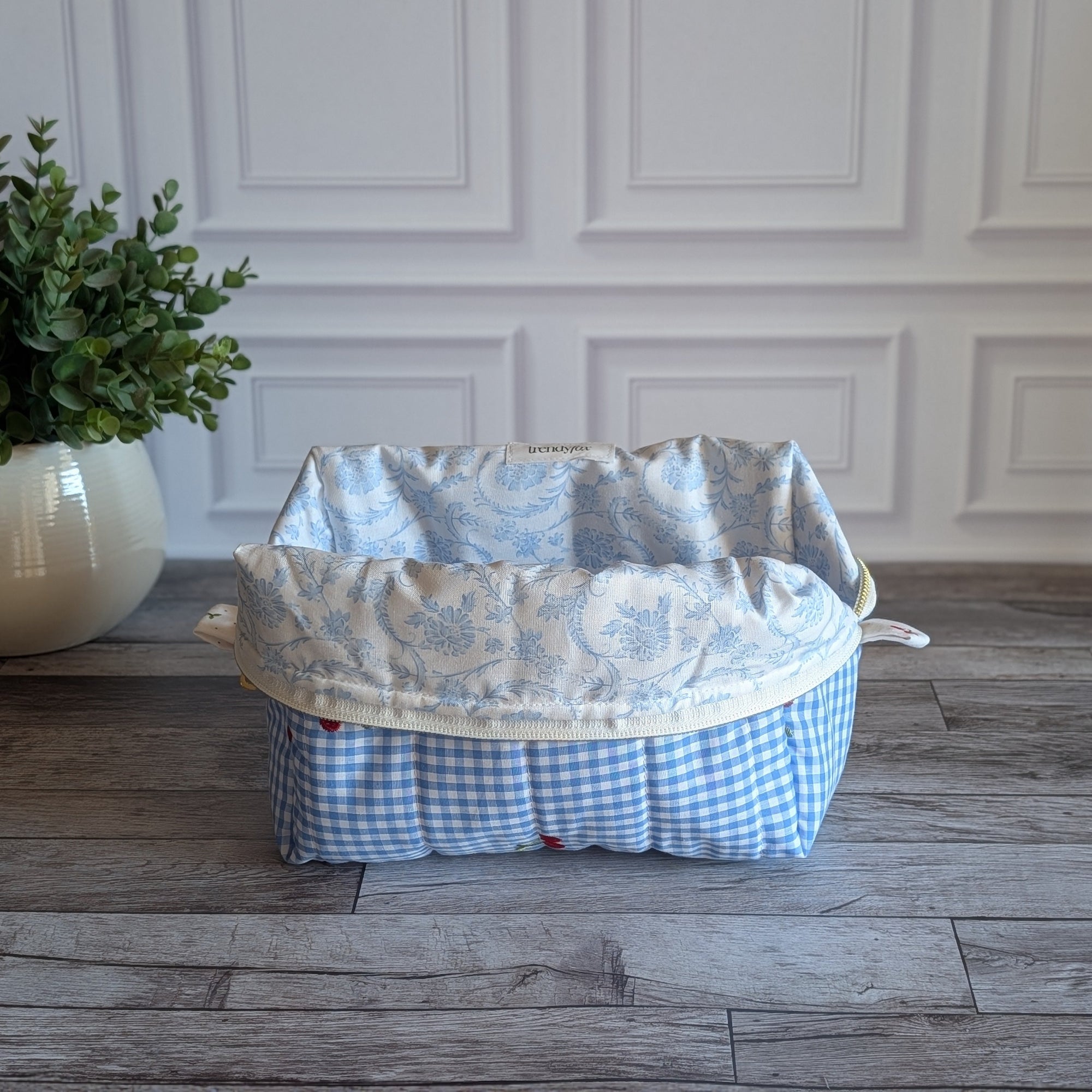 A light blue and white gingham toiletry bag with red cherries and green leaves embroidered on the front, sitting on a gray wood floor in front of a white paneled wall.