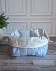 A light blue and white gingham toiletry bag with red cherries and green leaves embroidered on the front, sitting on a gray wood floor in front of a white paneled wall.
