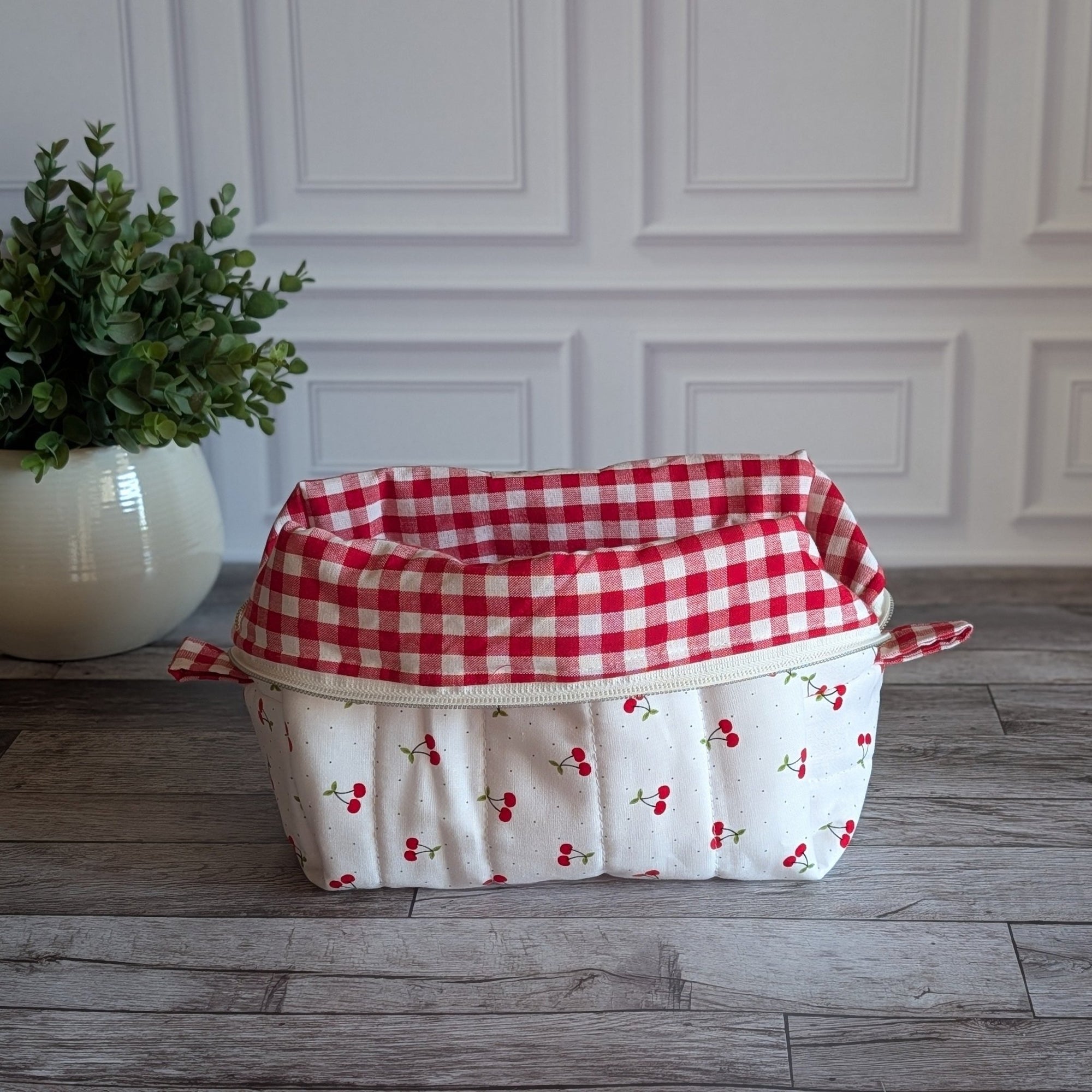 A quilted white cosmetic bag with a red cherry print and red gingham trim, sitting on a gray wood floor next to a potted green plant.