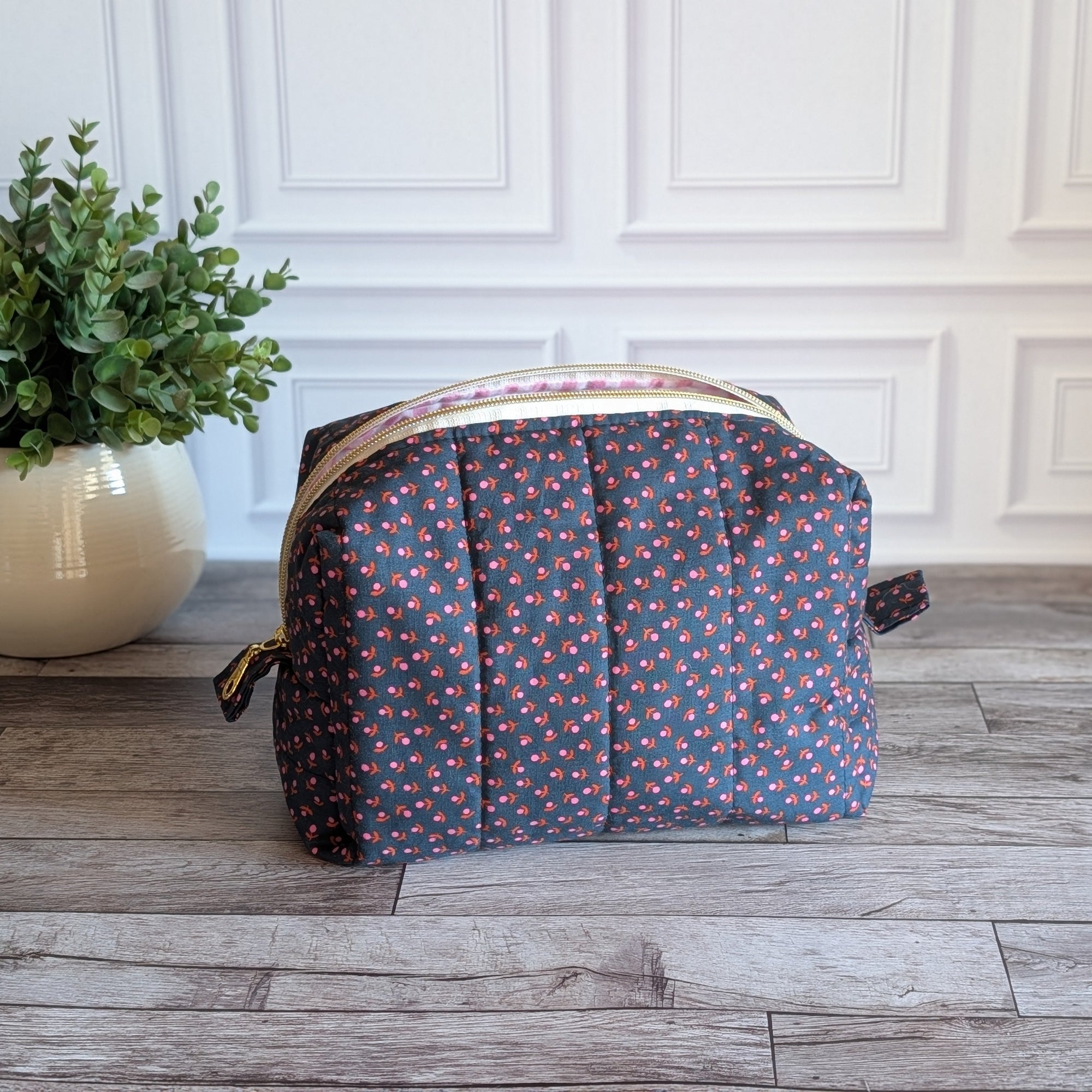 A quilted cosmetic pouch with a navy blue base and a small pink, orange, and yellow floral pattern, sitting on a gray wood floor in front of a white paneled wall with a potted plant on the left.