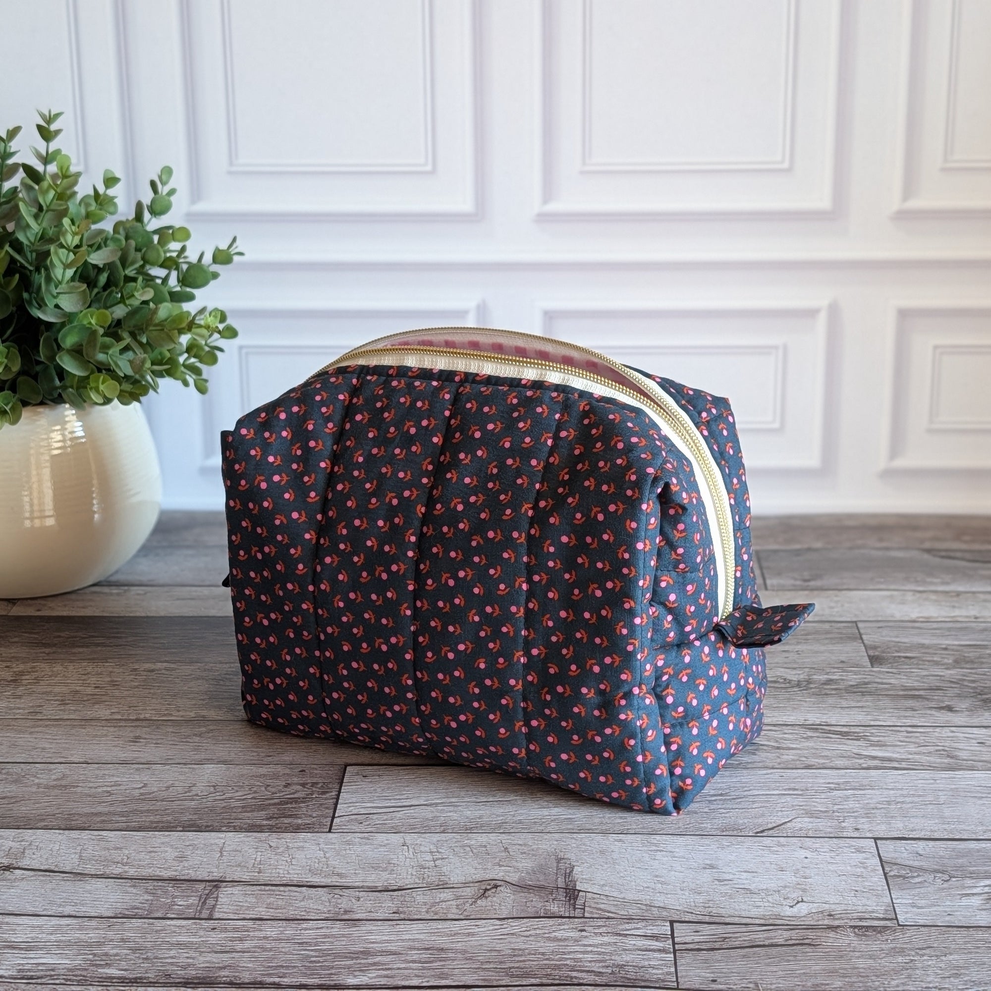 A toiletry bag with a dark navy blue base and a small pink and orange floral pattern, featuring a cream-colored zipper, set on a rustic wood floor next to a potted green plant.