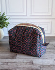 A toiletry bag with a dark navy blue base and a small pink and orange floral pattern, featuring a cream-colored zipper, set on a rustic wood floor next to a potted green plant.