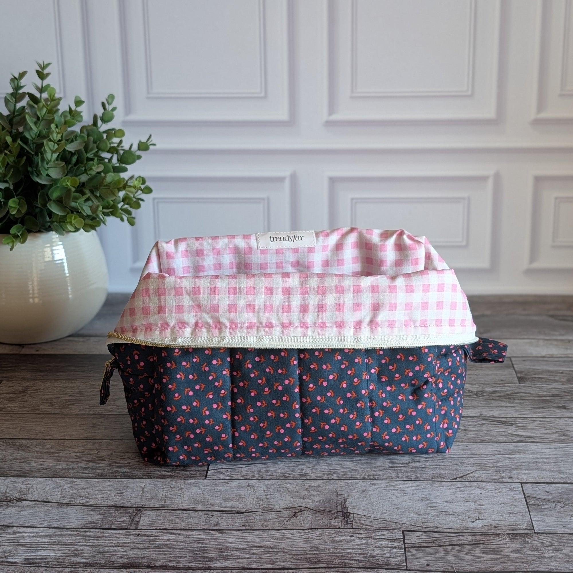 A quilted cosmetic pouch with a navy blue base and a small pink, orange, and yellow floral pattern, sitting on a gray wood floor in front of a white paneled wall with a potted plant on the left.