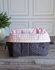 A quilted cosmetic pouch with a navy blue base and a small pink, orange, and yellow floral pattern, sitting on a gray wood floor in front of a white paneled wall with a potted plant on the left.
