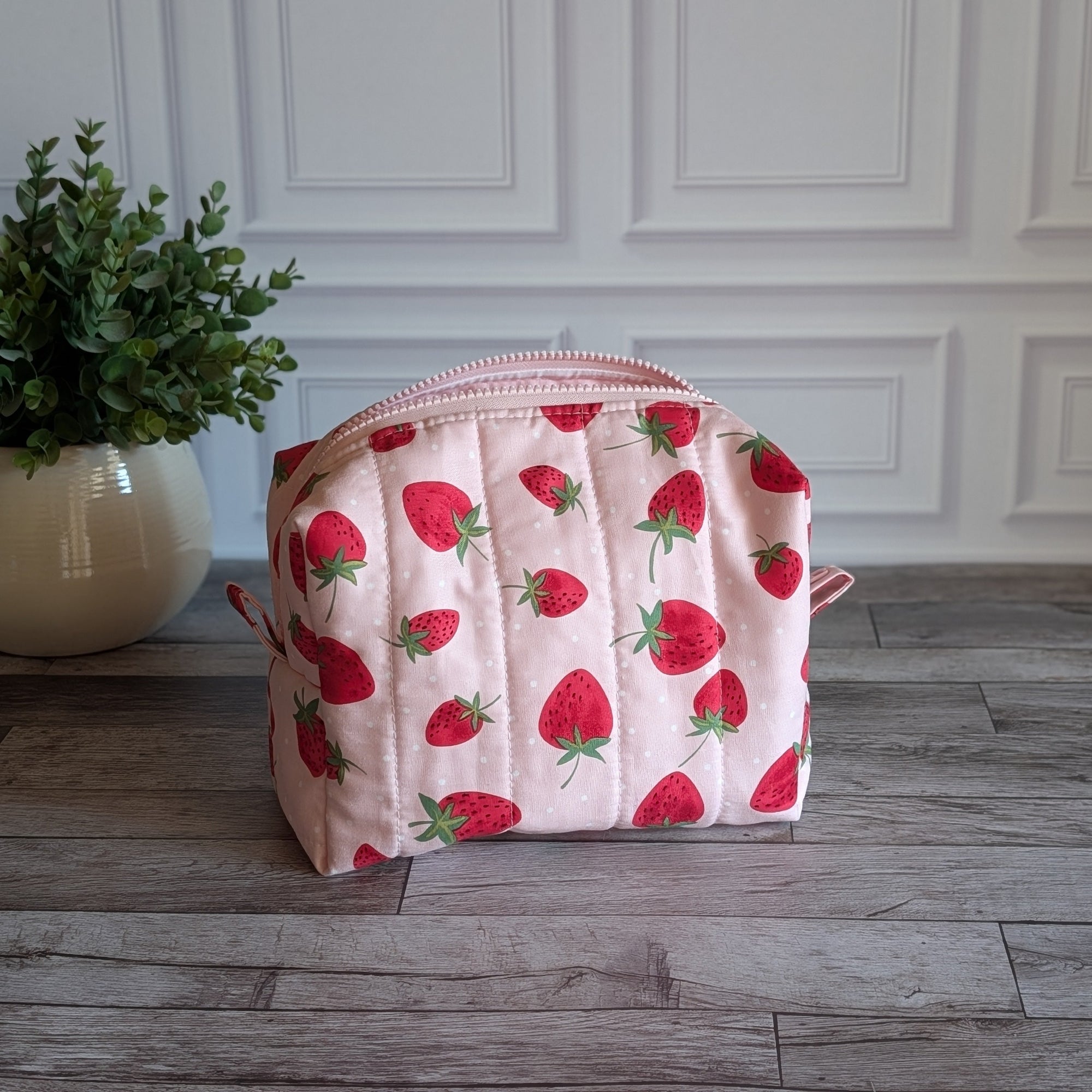 A quilted cosmetic pouch with a pink and white strawberry pattern, sitting on a gray wooden floor in front of a white paneled wall with a potted plant on the left.
