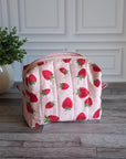 A quilted cosmetic pouch with a pink and white strawberry pattern, sitting on a gray wooden floor in front of a white paneled wall with a potted plant on the left.