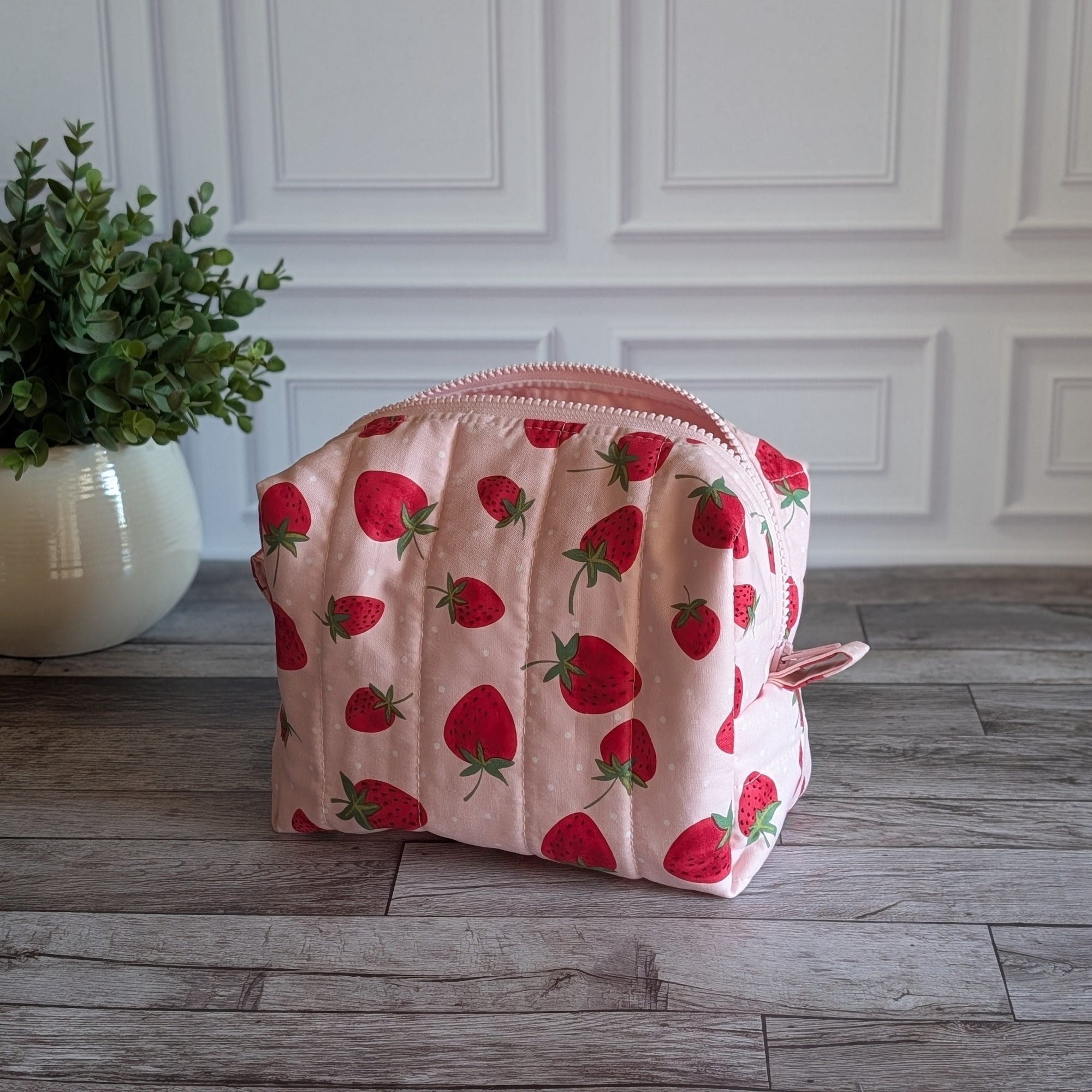 A quilted cosmetic pouch with a pink and red strawberry pattern, placed on a gray wood floor next to a potted green plant against a white paneled wall.