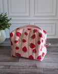A quilted cosmetic pouch with a pink and red strawberry pattern, placed on a gray wood floor next to a potted green plant against a white paneled wall.