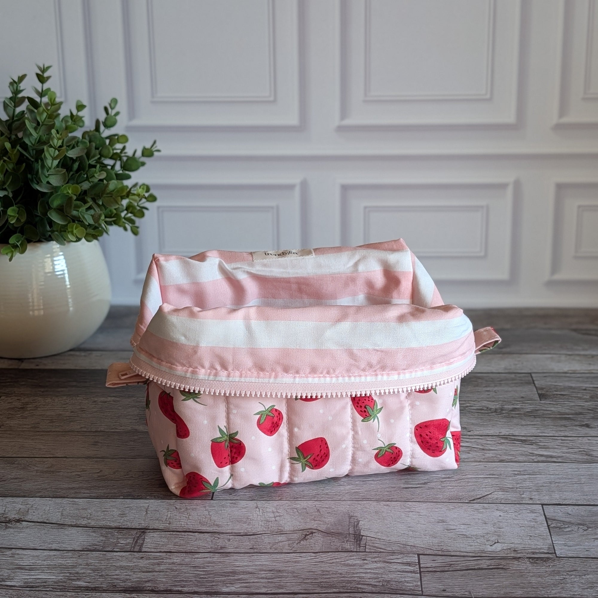 A quilted cosmetic pouch with a pink and white strawberry pattern, sitting on a gray wooden floor in front of a white paneled wall with a potted plant on the left.