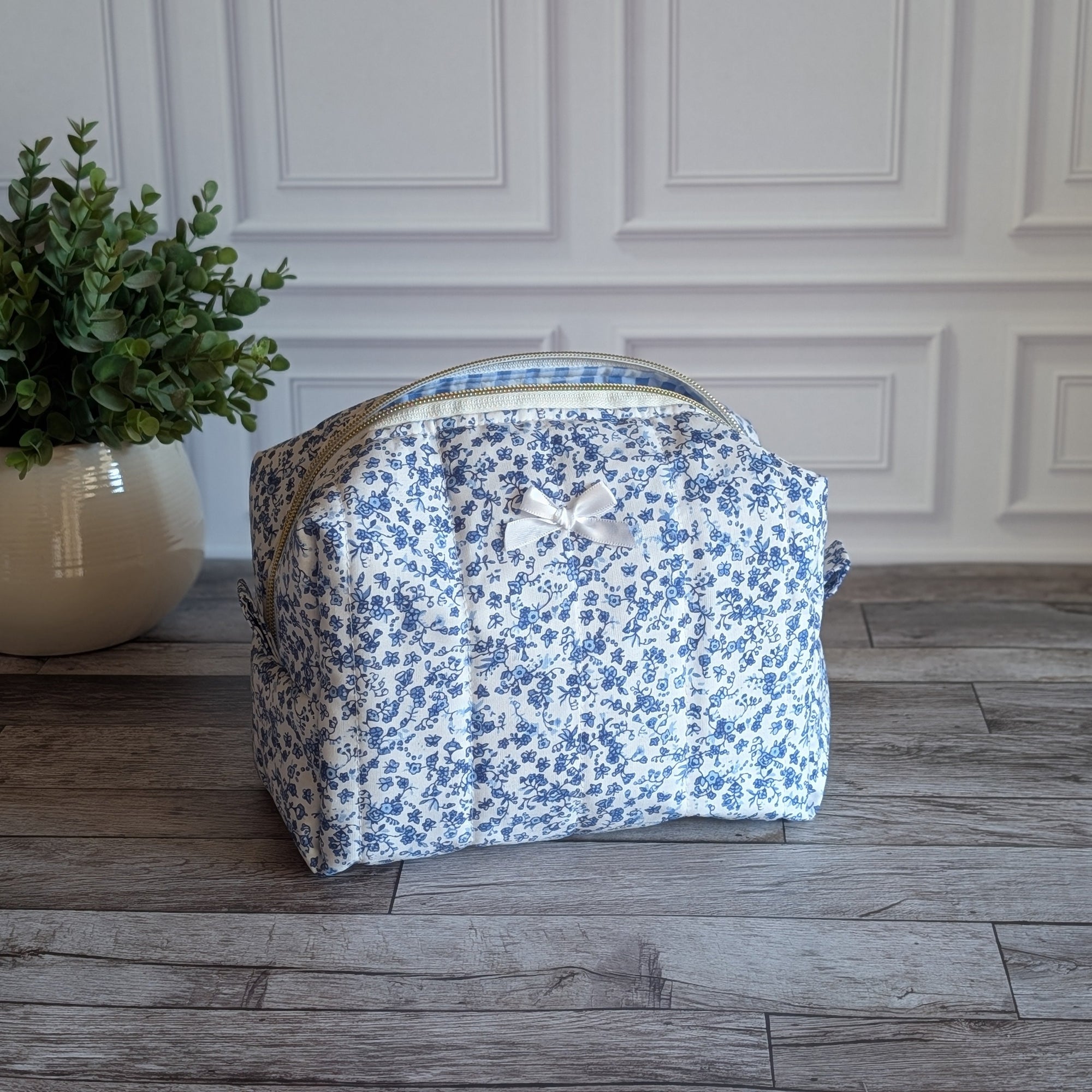 A fabric cosmetic bag with a blue and white floral print sits on a gray wood plank floor in front of a white paneled wall, next to a potted green plant.