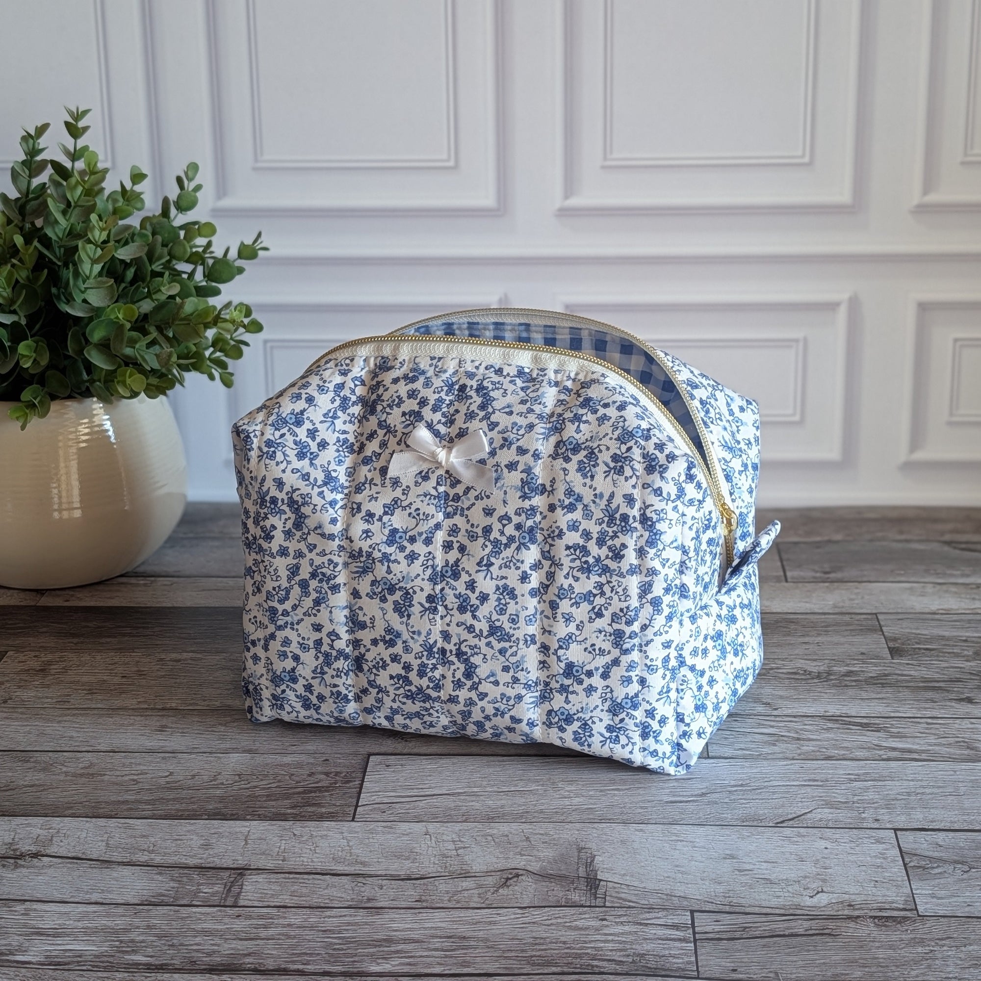 A fabric cosmetic bag with a blue and white floral print sits on a gray wood plank floor in front of a white paneled wall, next to a potted green plant.