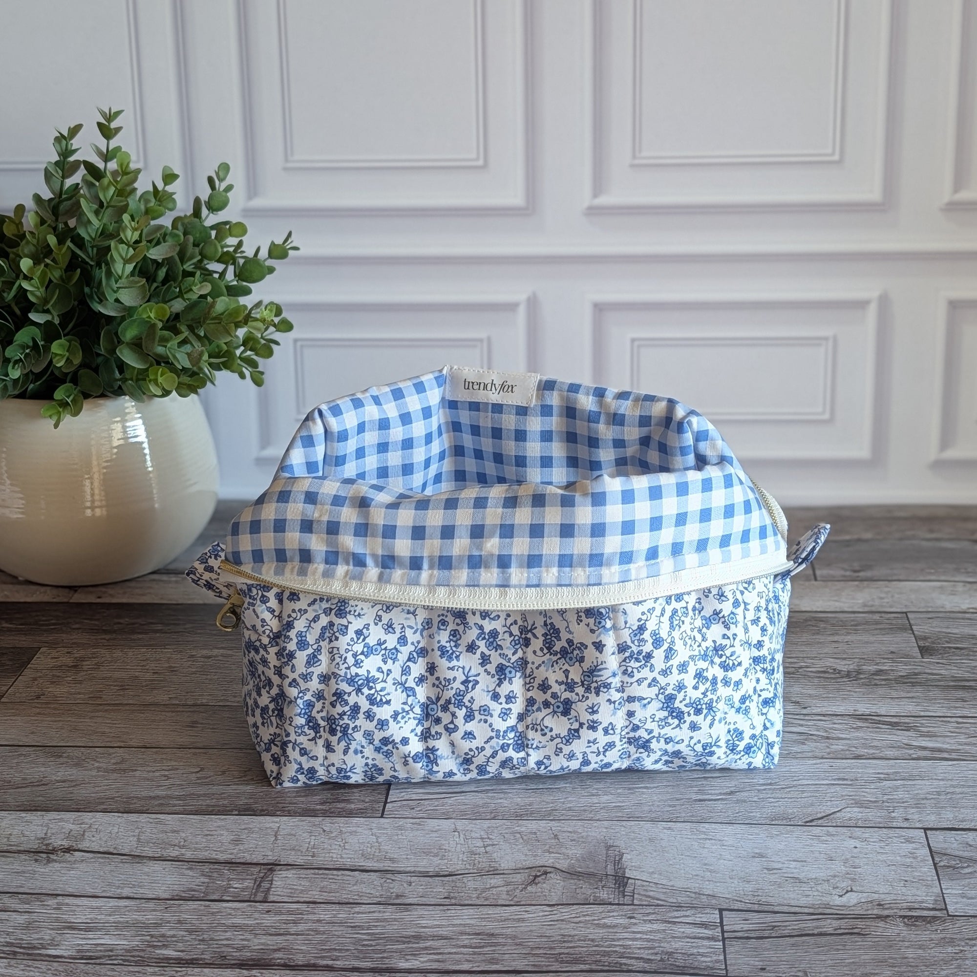 A fabric cosmetic bag with a blue and white floral print sits on a gray wood plank floor in front of a white paneled wall, next to a potted green plant.
