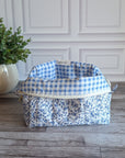 A fabric cosmetic bag with a blue and white floral print sits on a gray wood plank floor in front of a white paneled wall, next to a potted green plant.