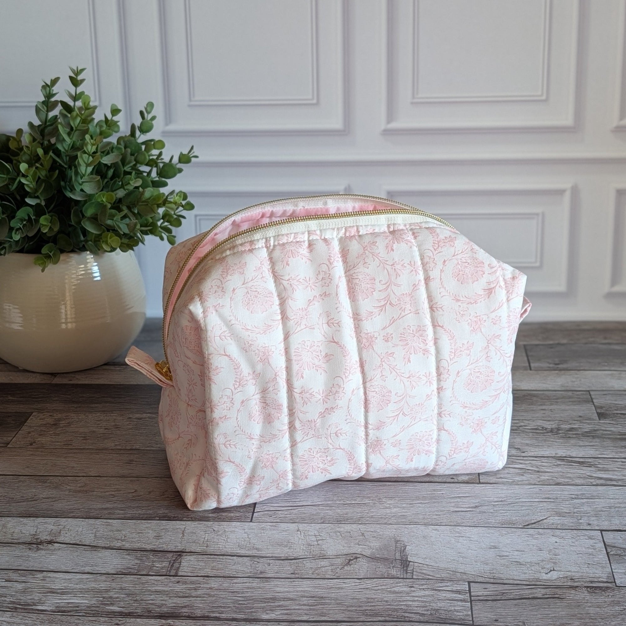 A light pink and white patterned fabric cosmetic pouch with a pleated design, sitting on a gray wood floor next to a potted green plant against a white paneled wall.