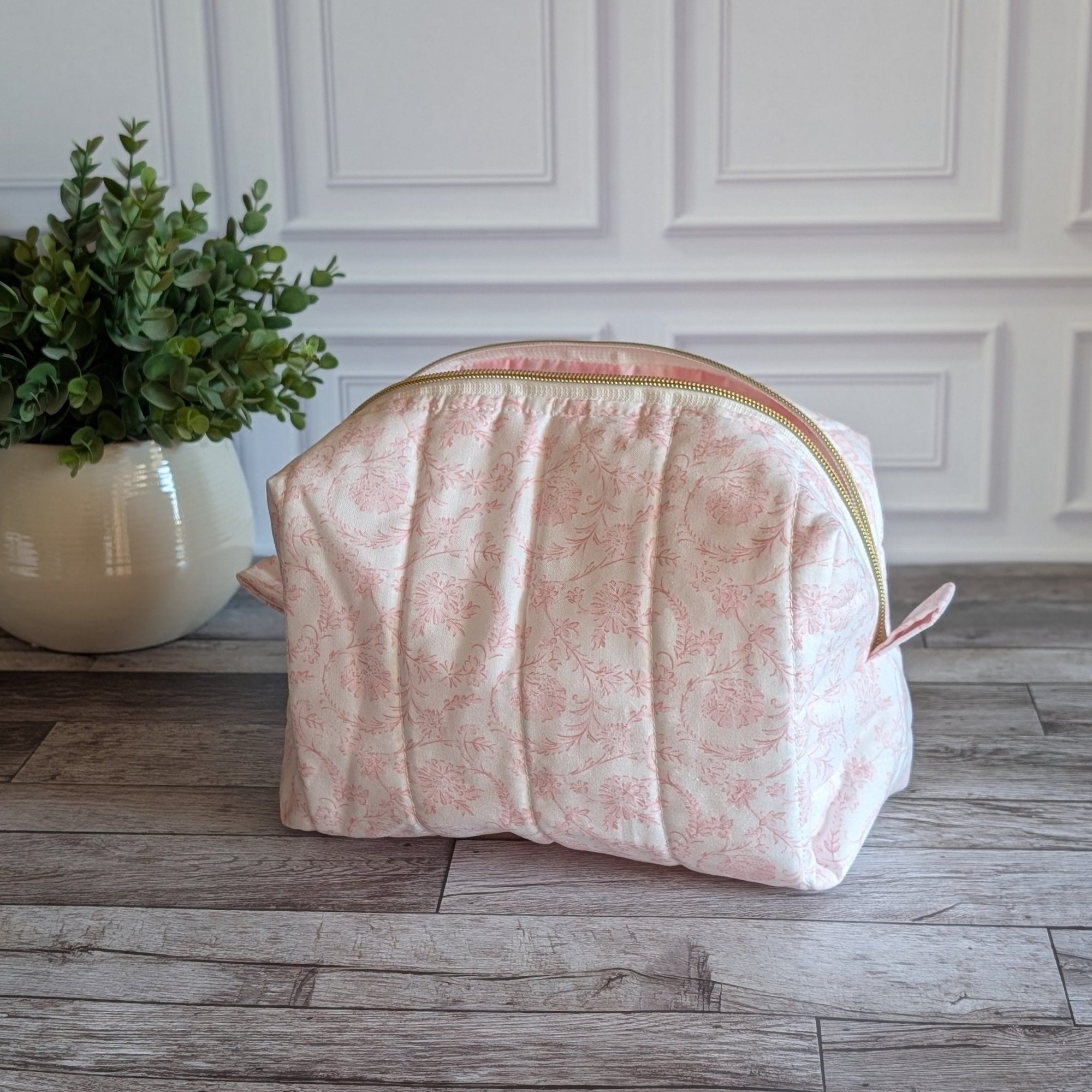 A light pink floral print zippered cosmetic pouch or case sits on a gray wood floor in front of a white paneled wall, with a potted green plant to the left.