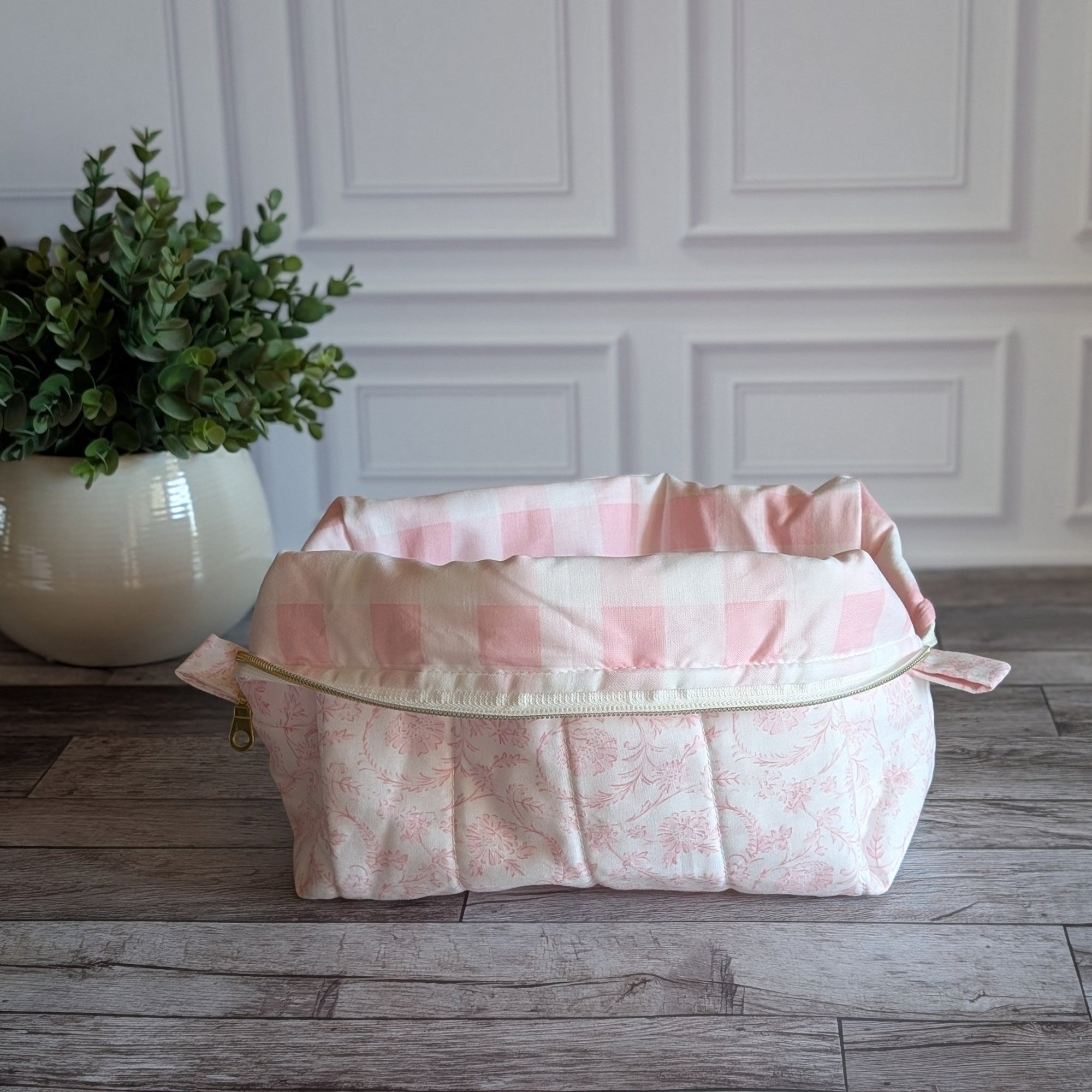 A pink and white checkered lining fabric quilted pouch, placed on a gray wood floor in front of a white paneled wall, with a potted green plant on the left.
