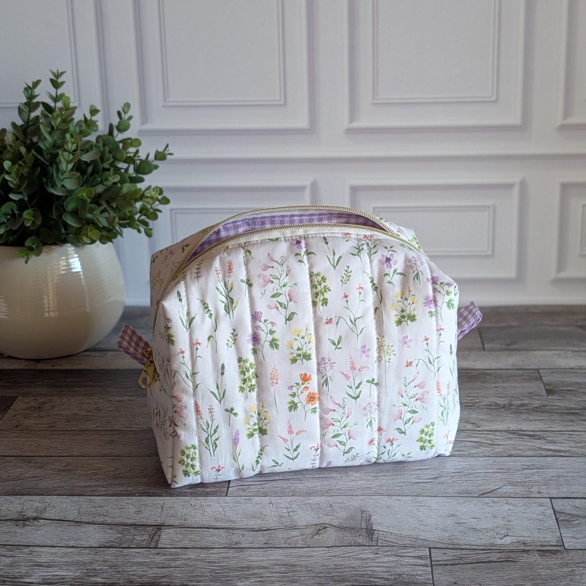 A quilted cosmetic bag with a pastel floral print and purple gingham trim, sitting on a gray wood floor next to a potted green plant against a white paneled wall.