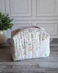 A quilted cosmetic bag with a pastel floral print and purple gingham trim, sitting on a gray wood floor next to a potted green plant against a white paneled wall.
