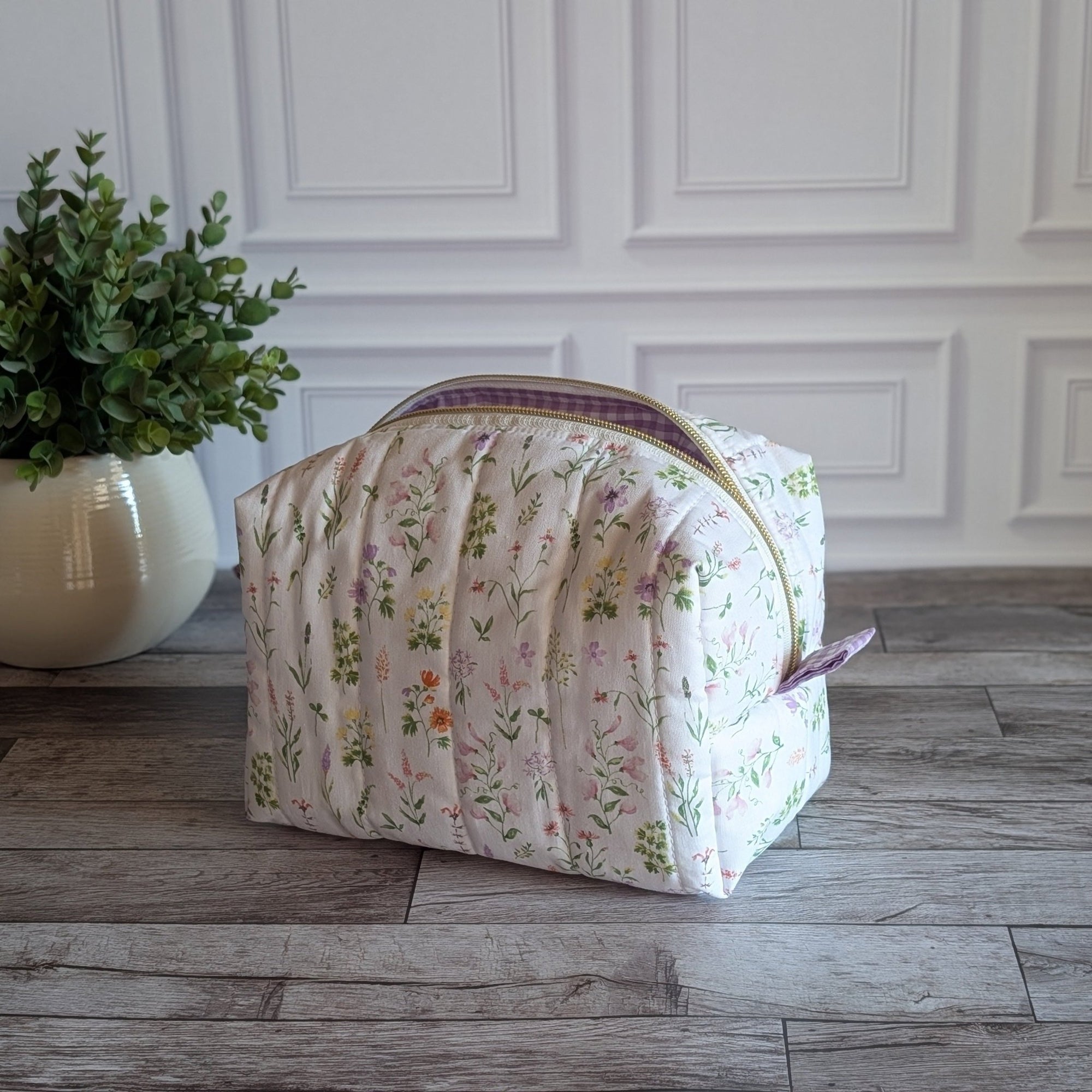 A quilted cosmetic bag with a pink and green floral print sits on a gray wood floor in front of a white paneled wall, next to a potted green plant.