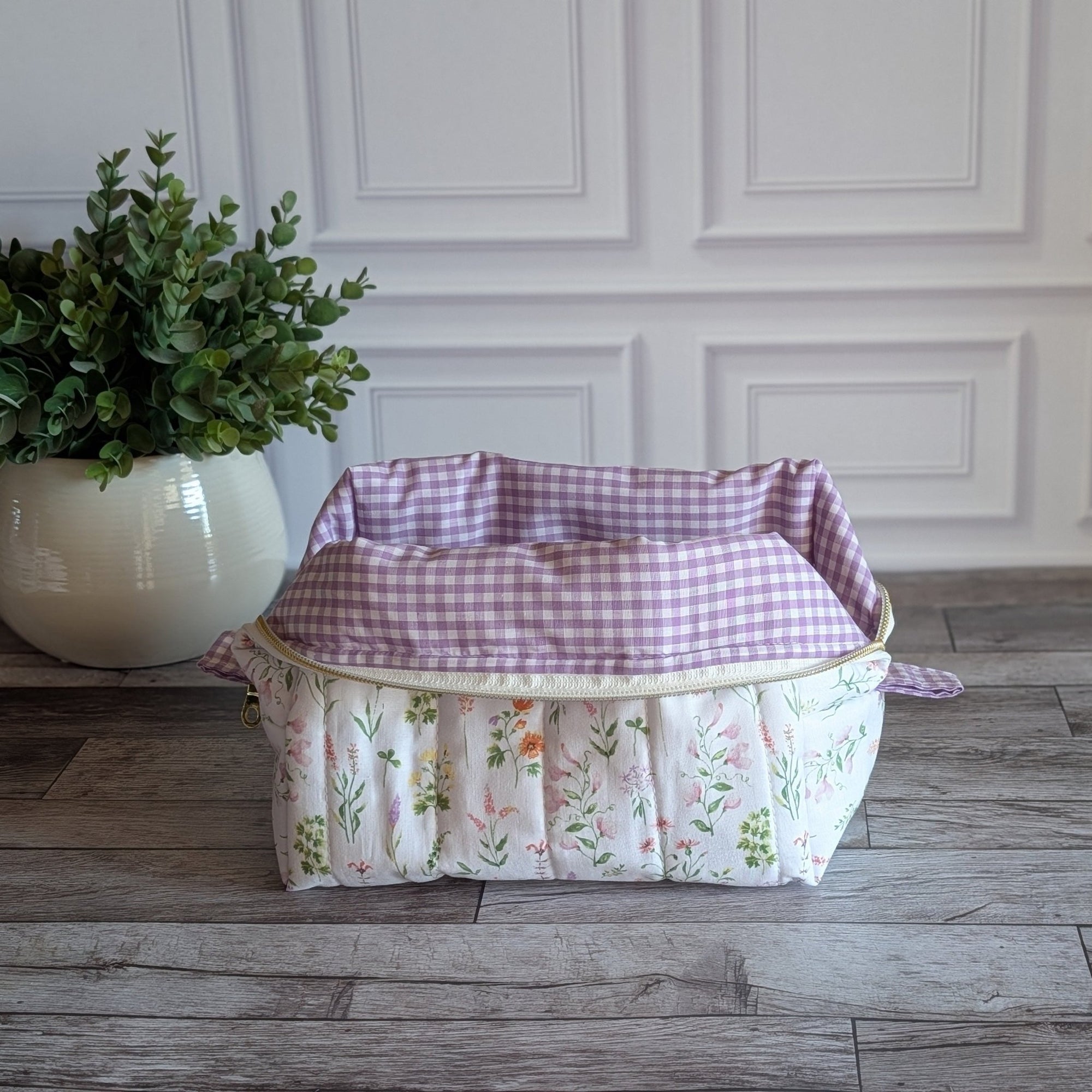A quilted cosmetic bag with a pink and green floral print sits on a gray wood floor in front of a white paneled wall, next to a potted green plant.