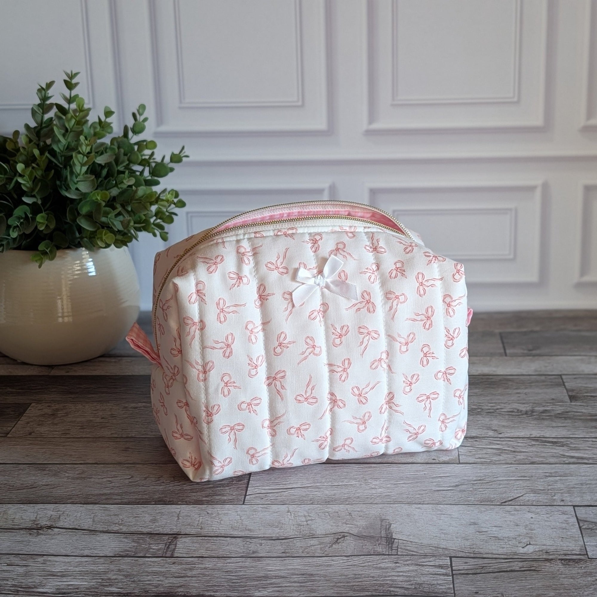 A quilted cosmetic bag with a pink and white bow pattern, sitting on a gray wood floor in front of a white paneled wall next to a potted green plant.