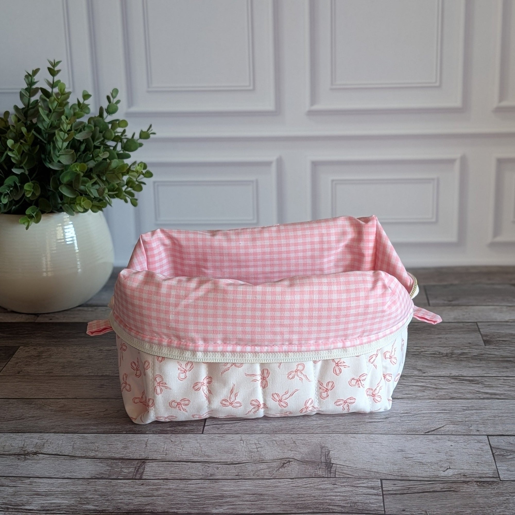 A quilted cosmetic bag with a pink and white bow pattern, sitting on a gray wood floor in front of a white paneled wall next to a potted green plant.