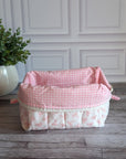A quilted cosmetic bag with a pink and white bow pattern, sitting on a gray wood floor in front of a white paneled wall next to a potted green plant.