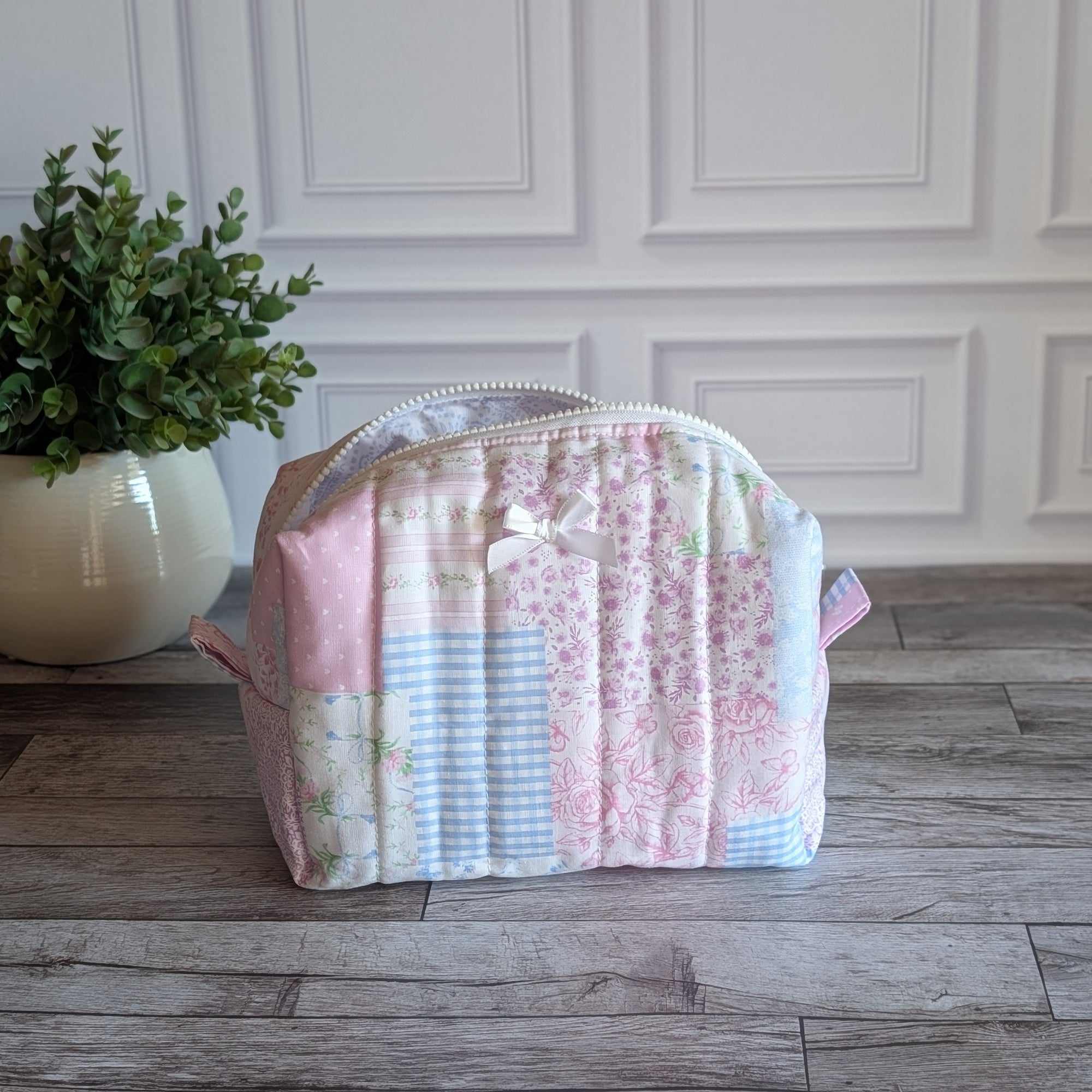 A patchwork quilted cosmetic bag in pastel pink, blue, and white fabrics, sitting on a gray wooden floor in front of a white paneled wall with a potted plant on the left.