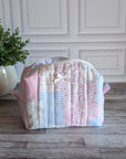 A patchwork quilted cosmetic bag in pastel pink, blue, and white fabrics, sitting on a gray wooden floor in front of a white paneled wall with a potted plant on the left.