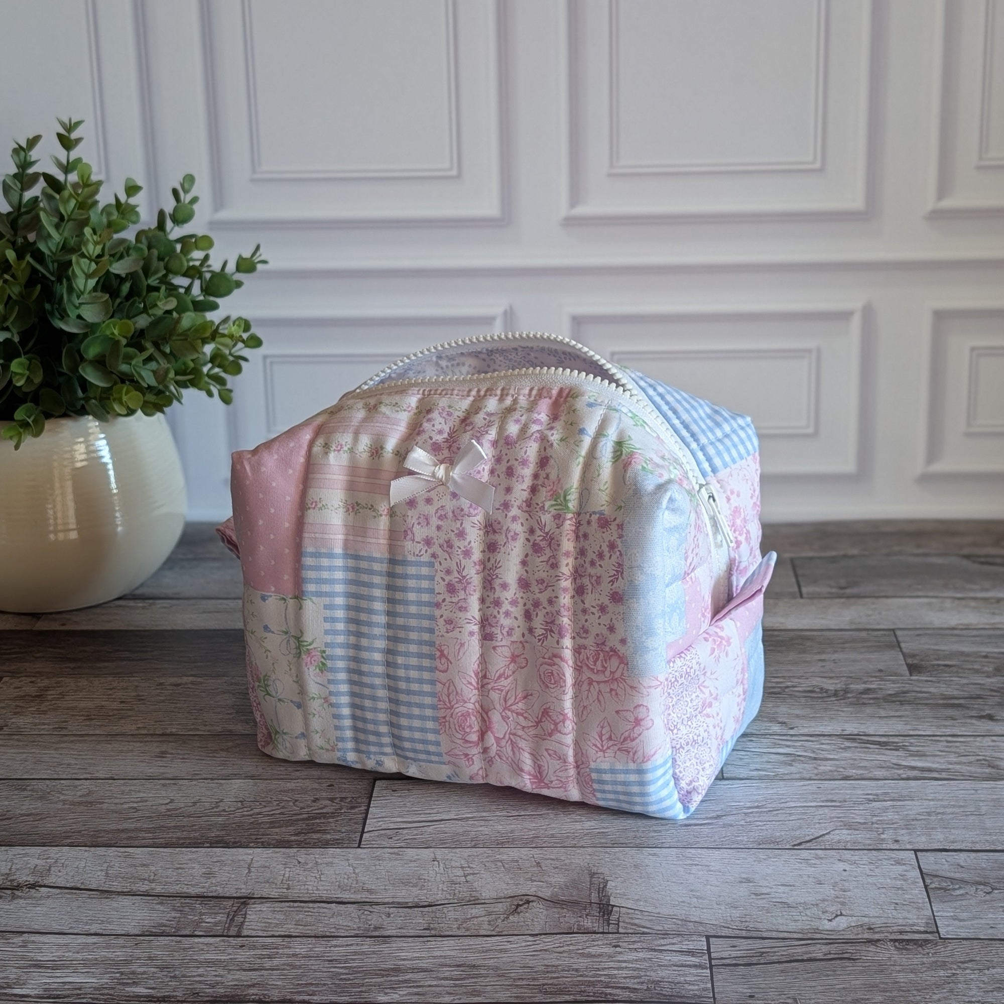 A patchwork quilted cosmetic bag in pastel pink, blue, and white fabrics, sitting on a gray wooden floor in front of a white paneled wall with a potted plant on the left.