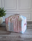 A patchwork quilted cosmetic bag in pastel pink, blue, and white fabrics, sitting on a gray wooden floor in front of a white paneled wall with a potted plant on the left.