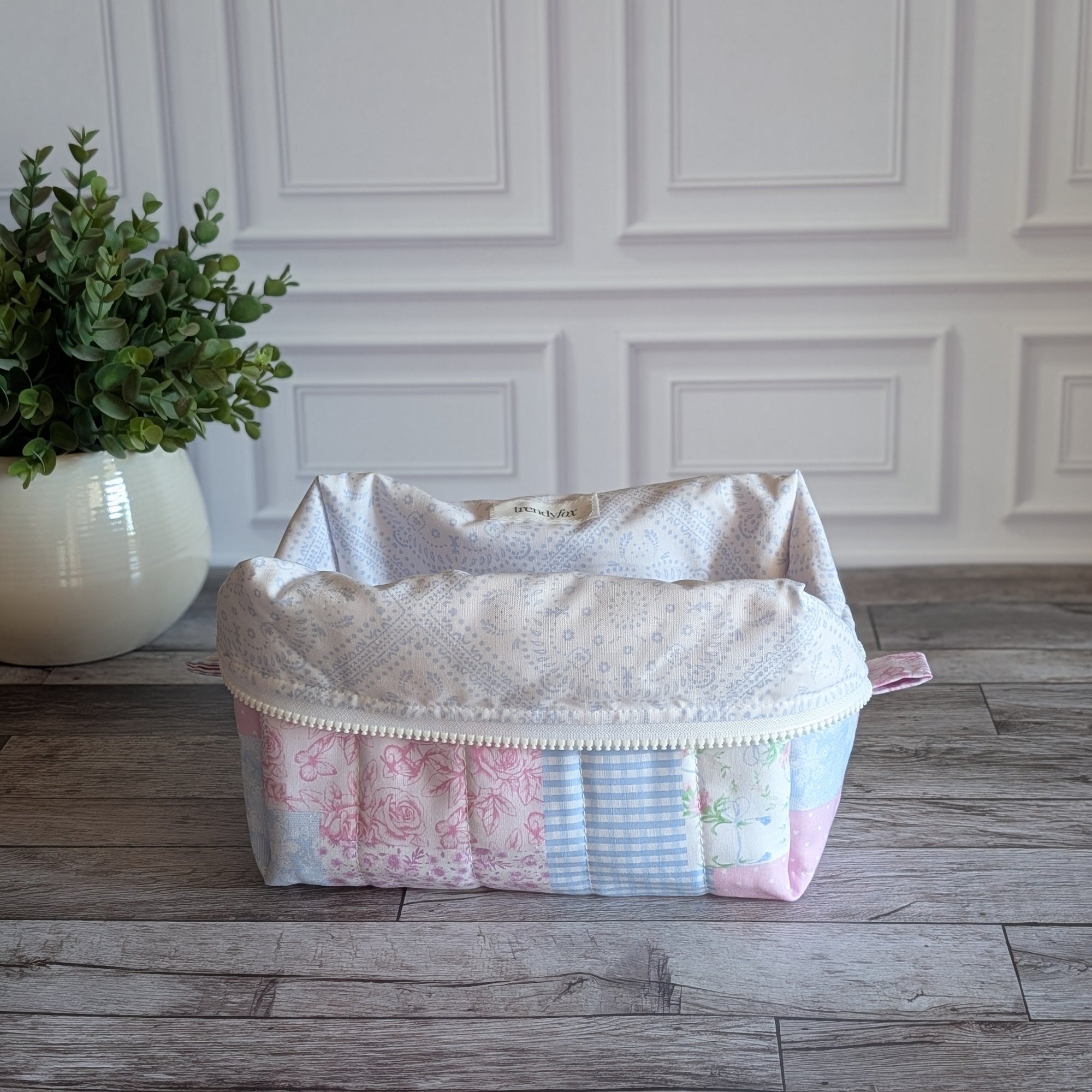 A patchwork quilted cosmetic bag in pastel pink, blue, and white fabrics, sitting on a gray wooden floor in front of a white paneled wall with a potted plant on the left.