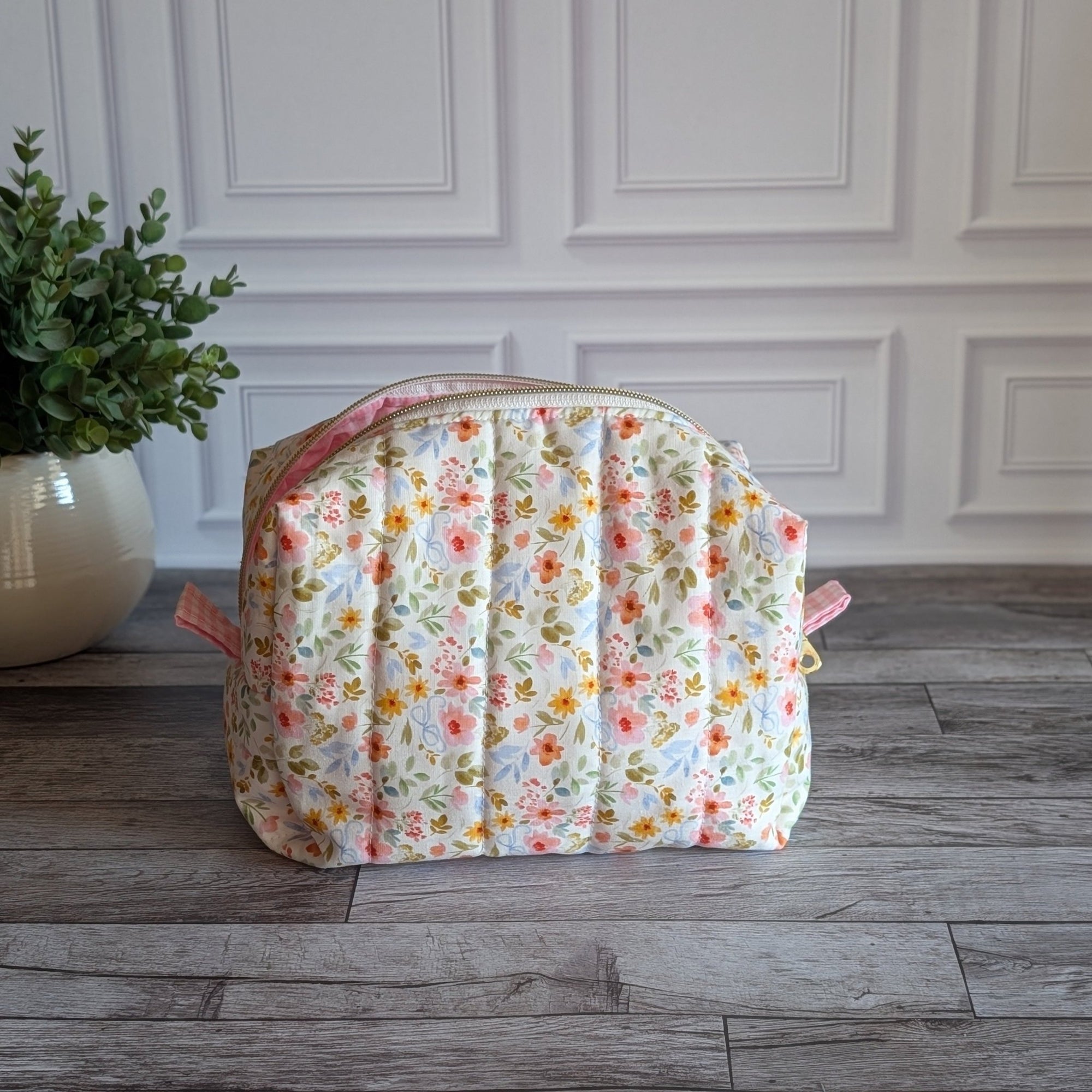 A quilted cosmetic pouch with a pink and yellow floral pattern on a white background, sitting on a gray wood floor next to a potted green plant.