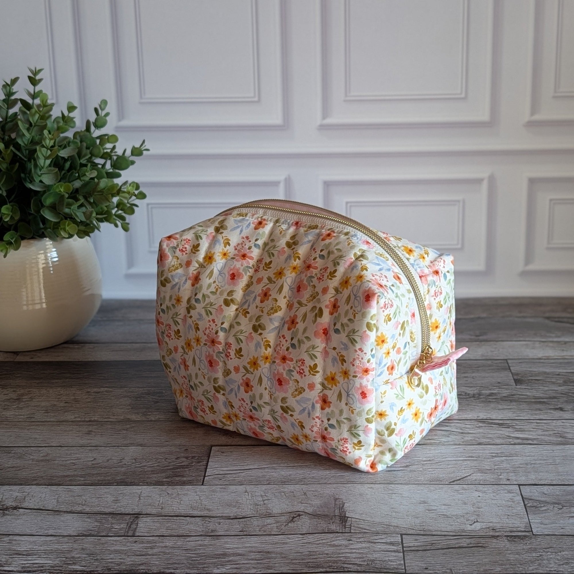 A fabric cosmetic pouch with a small multicolor floral print sits on a gray wood floor in front of a white paneled wall, next to a potted green plant.