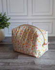 A fabric cosmetic pouch with a small multicolor floral print sits on a gray wood floor in front of a white paneled wall, next to a potted green plant.