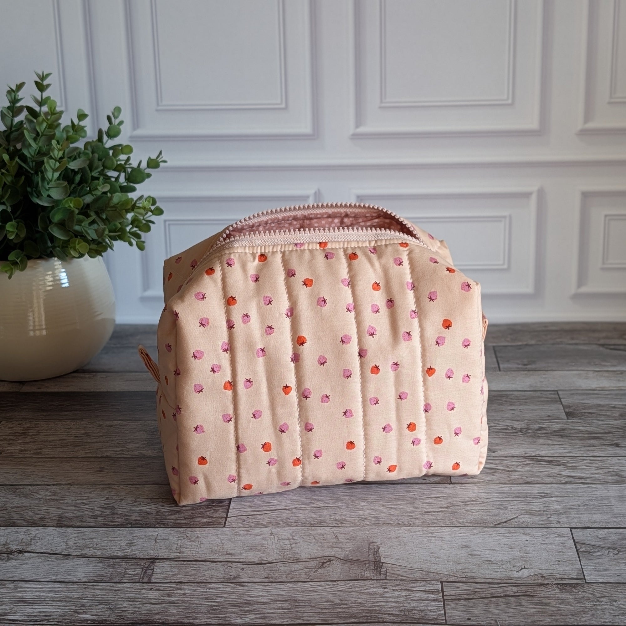 A quilted cosmetic pouch in a light peach-pink color with a pattern of small orange and pink strawberries, standing on a gray wood floor next to a potted green plant.
