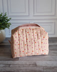 A quilted cosmetic pouch in a light peach-pink color with a pattern of small orange and pink strawberries, standing on a gray wood floor next to a potted green plant.