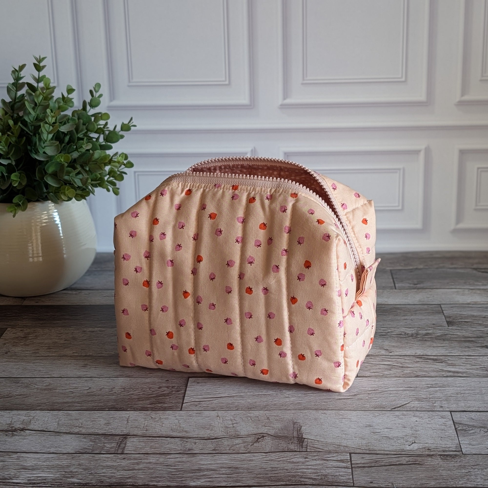 A quilted cosmetic pouch in a light peach-pink color with a pattern of small orange and pink strawberries, standing on a gray wood floor next to a potted green plant.